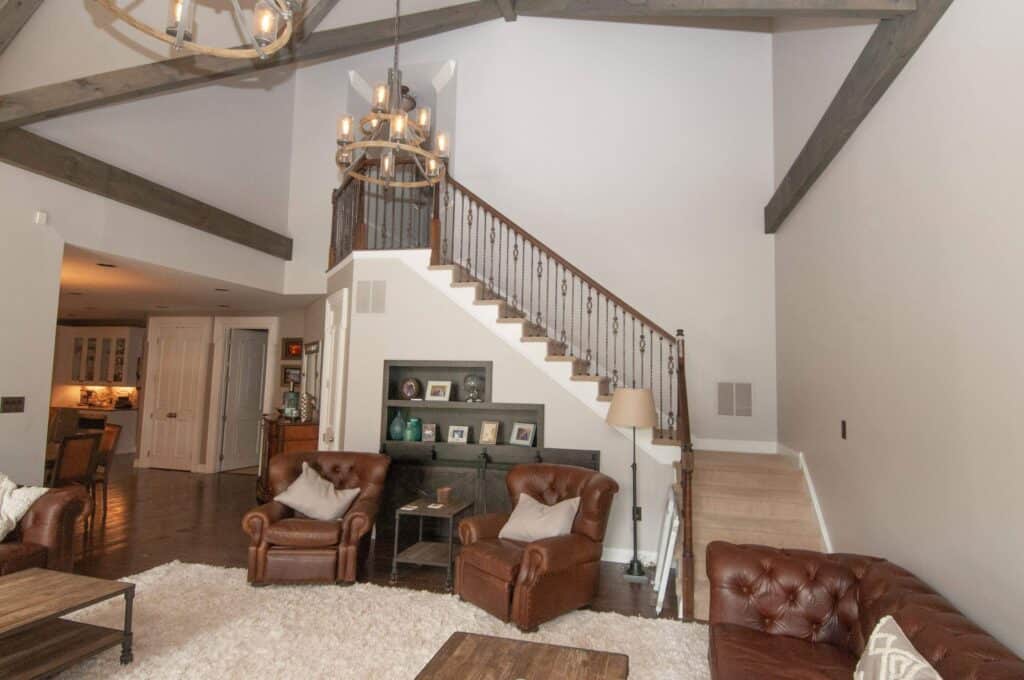 A spacious living room with high ceilings, exposed wooden beams, leather armchairs, a staircase with iron railings, a chandelier, wooden furniture, and neutral-colored walls and carpet.