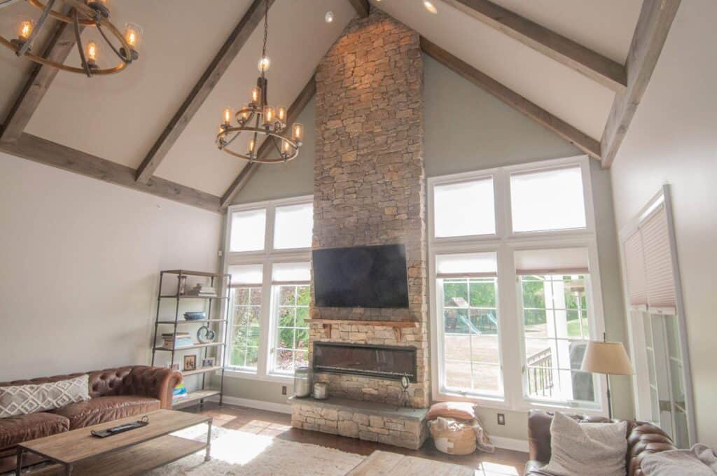 A bright living room with high vaulted ceilings, a tall stone fireplace, a wall-mounted TV, large windows, two brown leather couches, a wooden coffee table, and rustic chandeliers hanging from exposed beams.