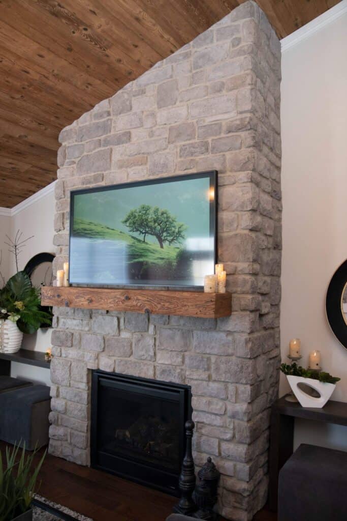 A stone fireplace with a wooden mantel displays lit candles and a framed landscape painting of a tree. Decorative items and plants are arranged on side tables against a wood-paneled ceiling.