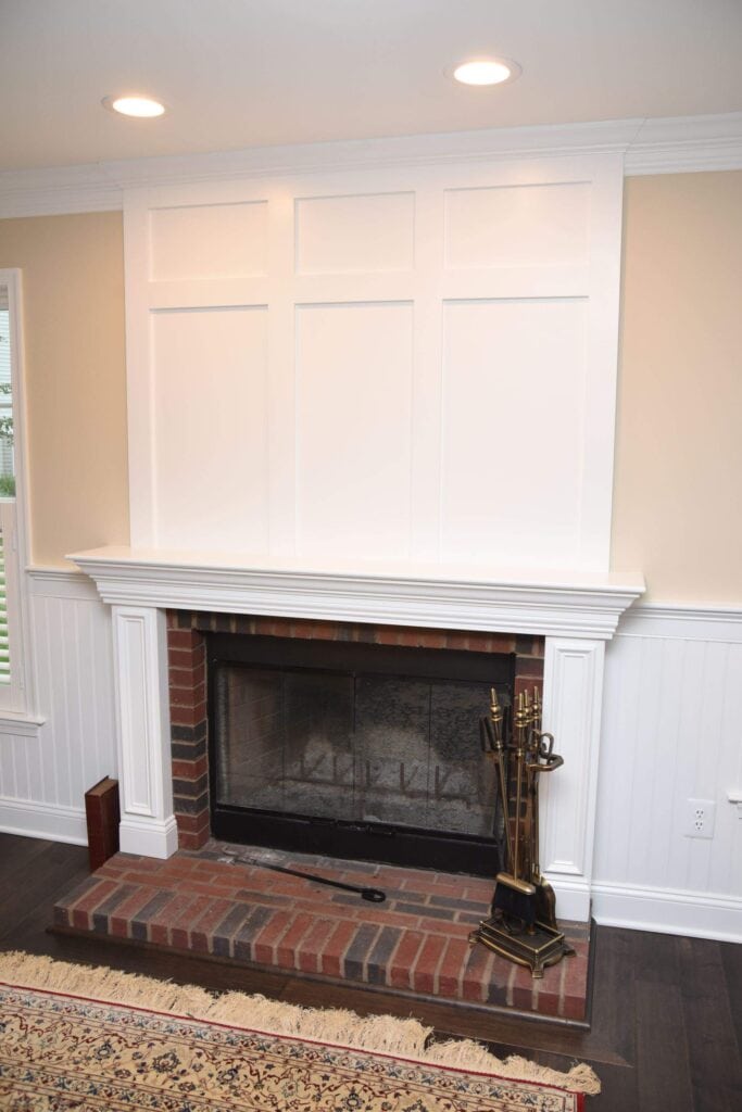 A white fireplace with a brick hearth and mantel, panel detailing above, and fireplace tools to the right, set against a beige wall with wainscoting and dark wood floors. A patterned rug is partially visible in front.
