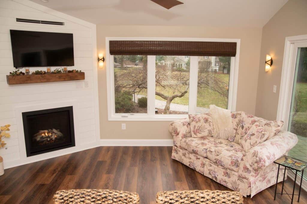 A cozy living room with a floral loveseat, a window overlooking a yard, a fireplace below a mounted TV, and warm wood floors. Wall sconces and a ceiling fan add to the inviting atmosphere.