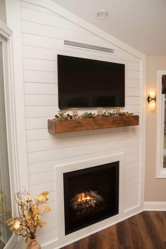 A modern living room features a white shiplap wall with a mounted flat-screen TV above a wooden mantel decorated with candles and foliage. Below the mantel, a lit fireplace creates a cozy atmosphere.