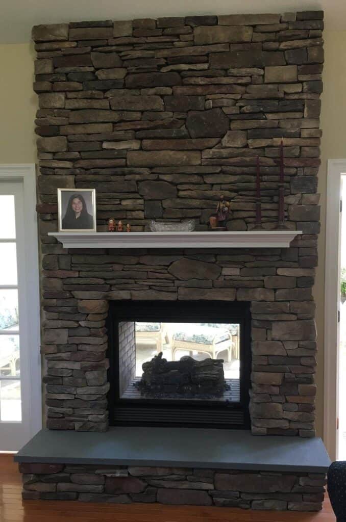 A stone fireplace with a double-sided firebox, featuring a white mantel holding a framed photo, glass dish, figurines, and two tall red candles. Sunlit rooms are visible through glass doors on either side.