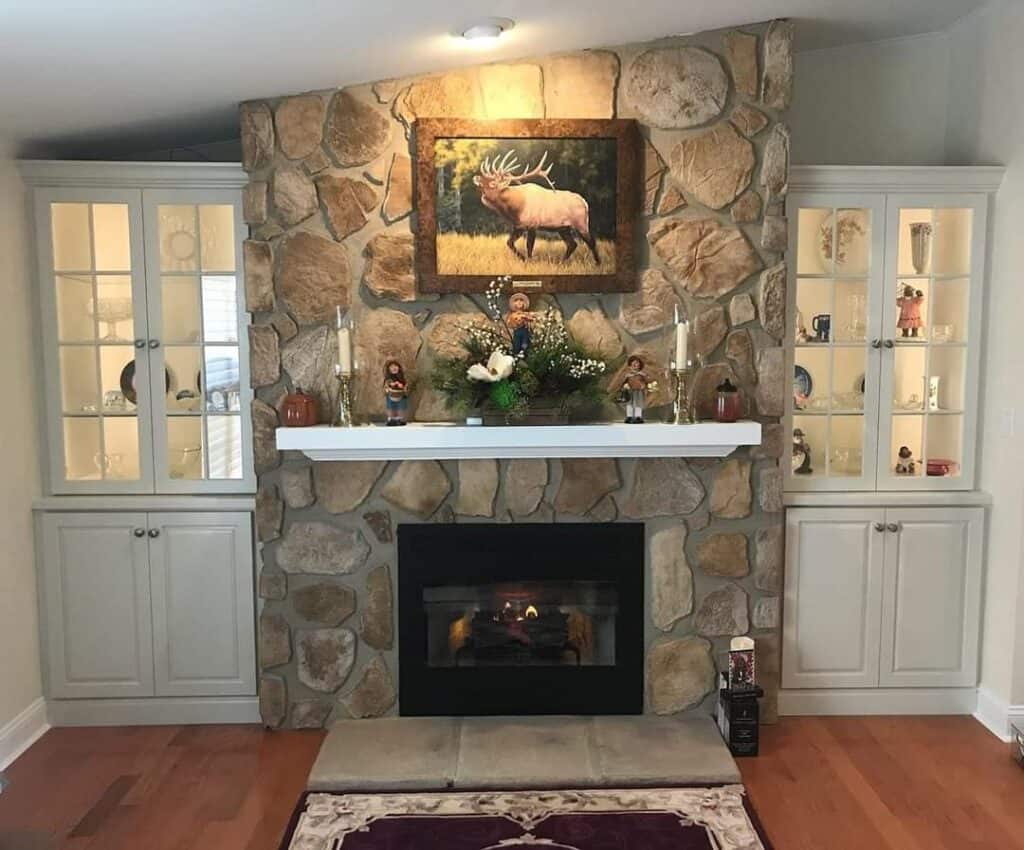 A stone fireplace with a white mantle displays candles, figurines, and flowers. Built-in cabinets with glass doors flank the fireplace. A painting of an elk hangs above, and a rug and hearth sit in front.