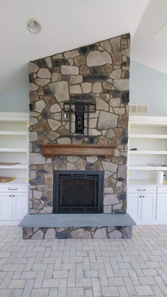 Stone fireplace with a wooden mantel, a black metal mount above for a TV, and built-in white shelves and cabinets on both sides. The floor is tiled in a light, rectangular pattern.