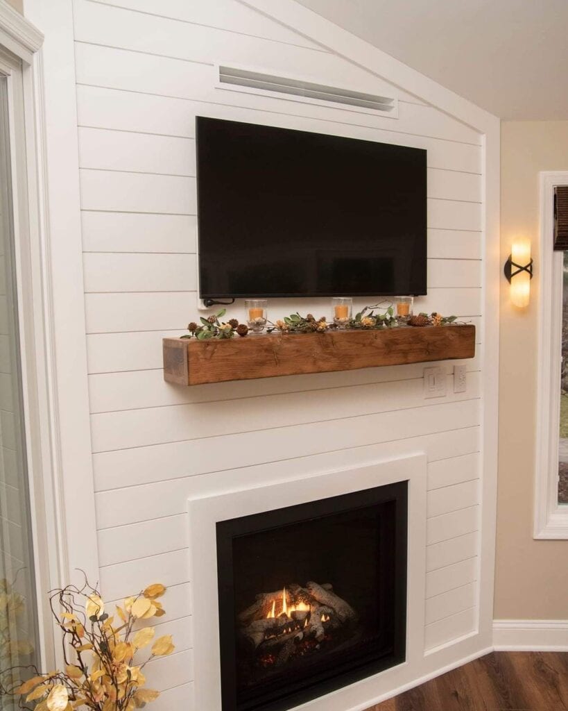 A modern living room corner features a black flat-screen TV mounted above a wooden mantel with fall decor and candles. Below, a fireplace with a glass front emits a warm glow. White shiplap walls and dark wood floors complete the space.