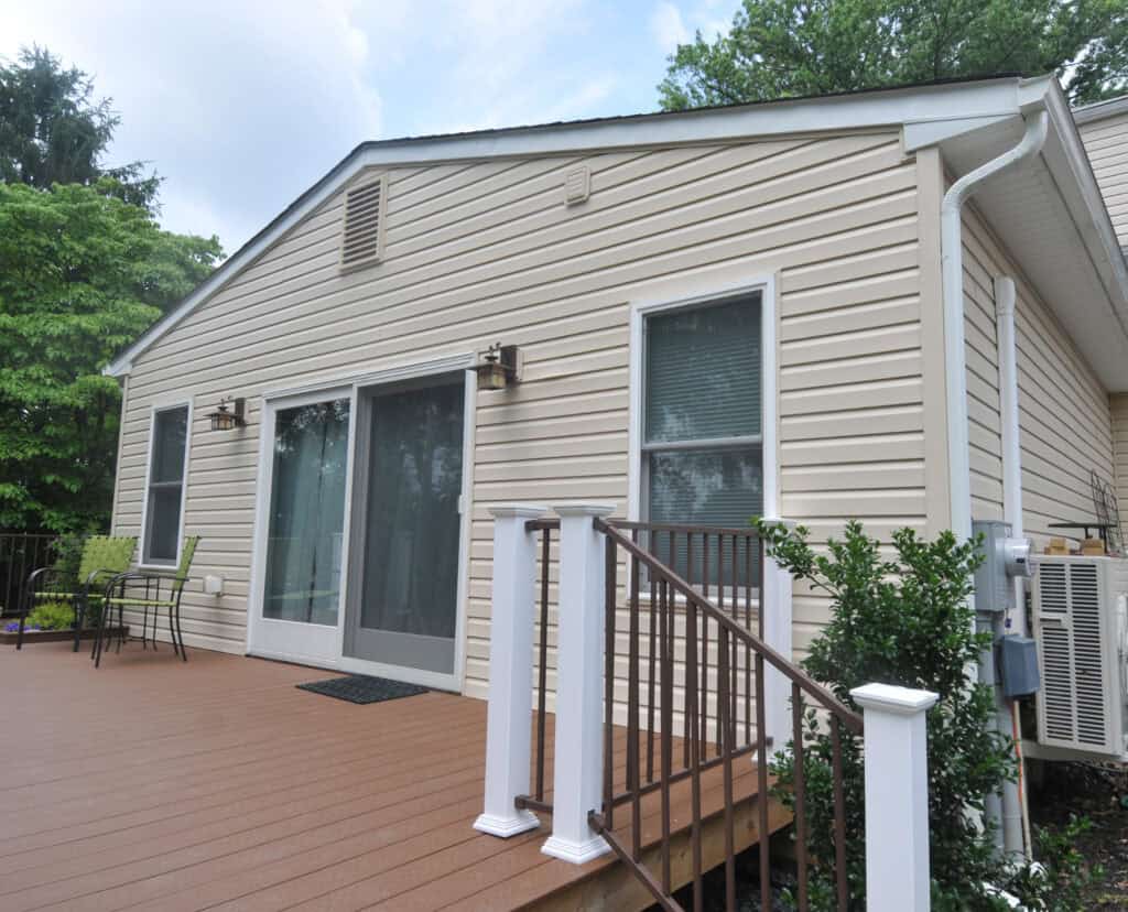 A beige house with white trim features a sliding glass door and windows opening onto a wooden deck with a small outdoor table and two chairs. Stairs with a railing lead down from the deck, surrounded by greenery.