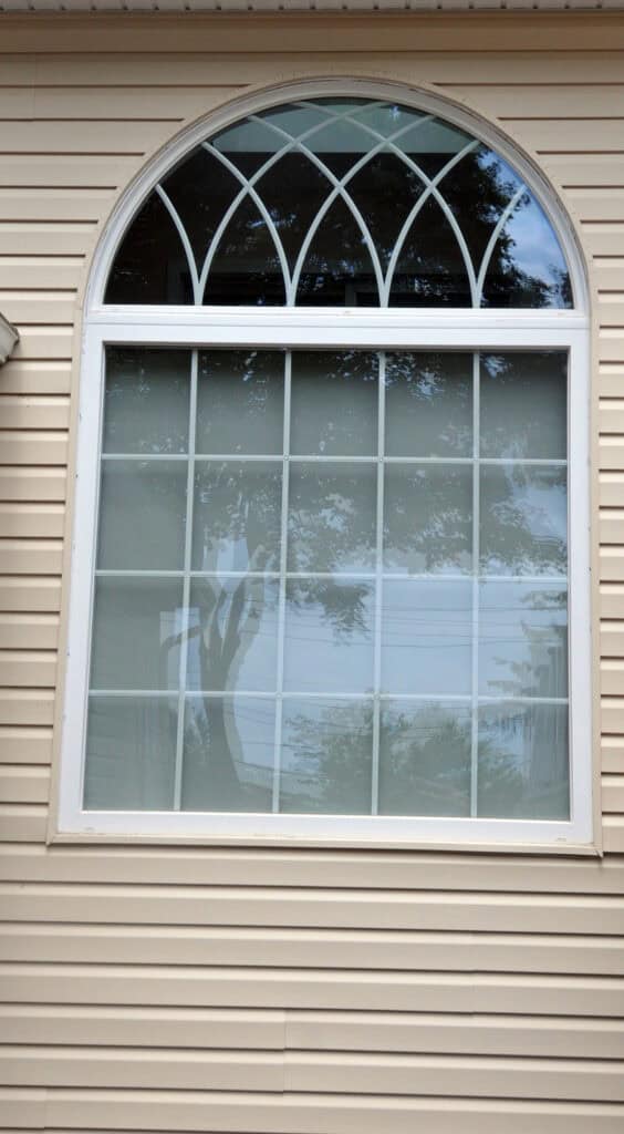 A large window with a rectangular lower section and an arched upper section featuring decorative white grid patterns, set in beige siding, reflecting trees and sky.