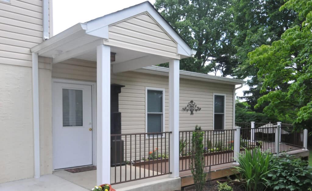 A beige house with white trim features a small covered porch, a white door, metal railing, and a ramp leading up to the entrance, surrounded by green trees and plants.