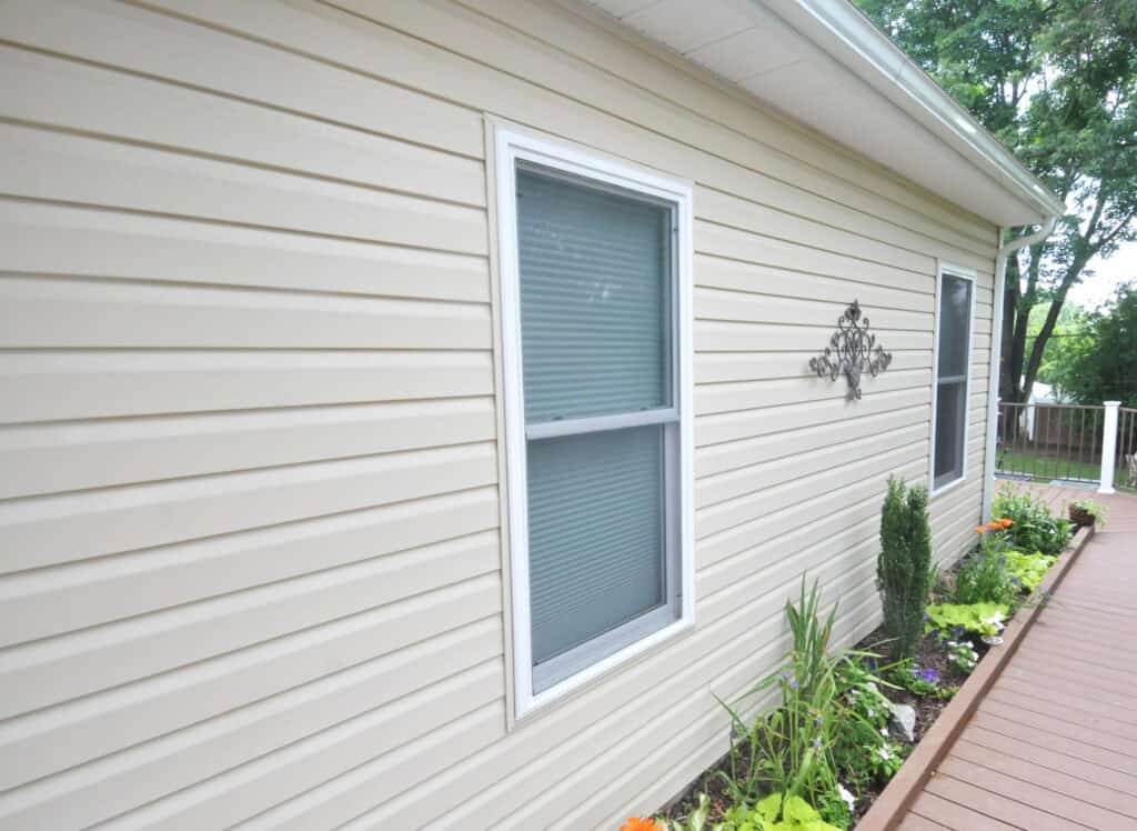 A beige vinyl-sided house with a white-framed window, a decorative metal wall hanging, and a garden bed with green plants and orange flowers next to a wooden walkway.