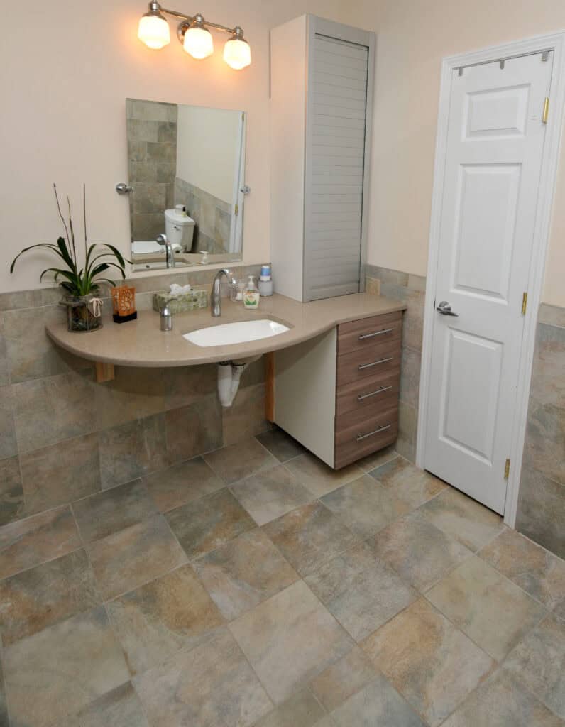 Modern bathroom with beige and gray tiled floor and walls, a countertop with sink, mirror, three-light fixture, cabinet with drawers, potted plant, and a white door on the right.