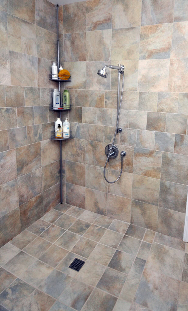 Walk-in shower with beige and gray tiles, a silver adjustable showerhead, corner shelves holding toiletries, and a square floor drain. There is no shower door or curtain visible.