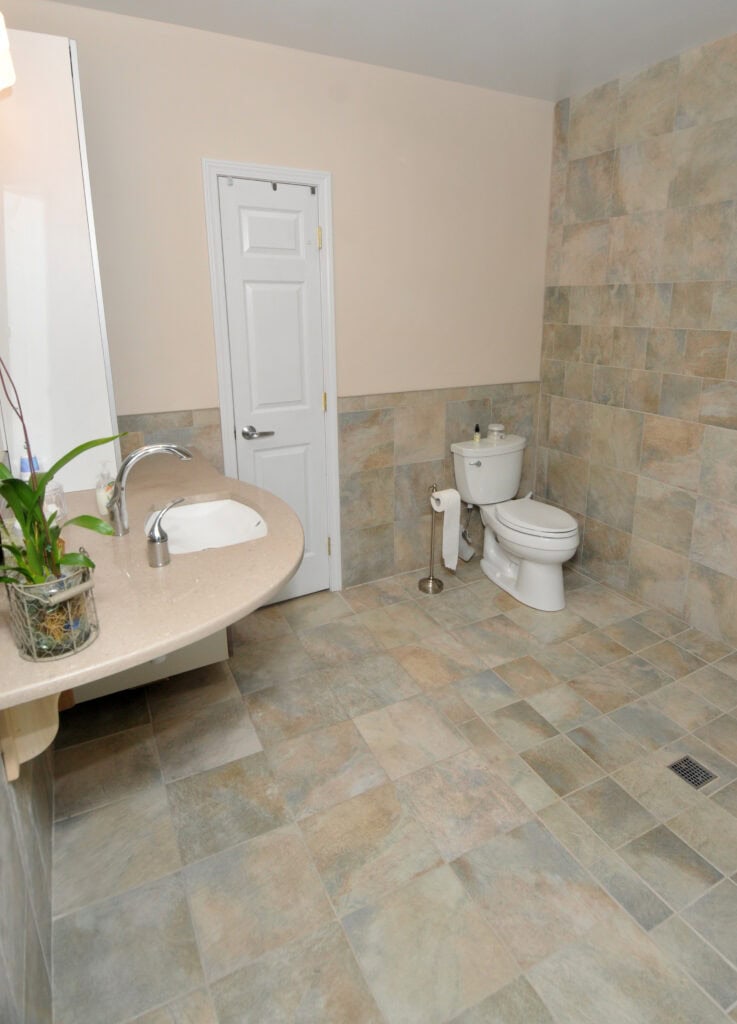 A spacious bathroom with tiled walls and floor, a white toilet, a wall-mounted sink, a mirror, a small potted plant on the counter, and a white door in the back wall.