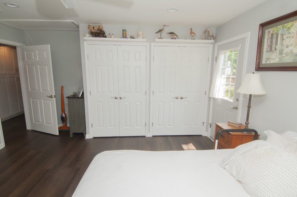A bright bedroom with white walls, dark wood floors, a neatly made bed, and several white closet doors. There are decorative items on a small cabinet, a floor lamp, and a window letting in natural light.