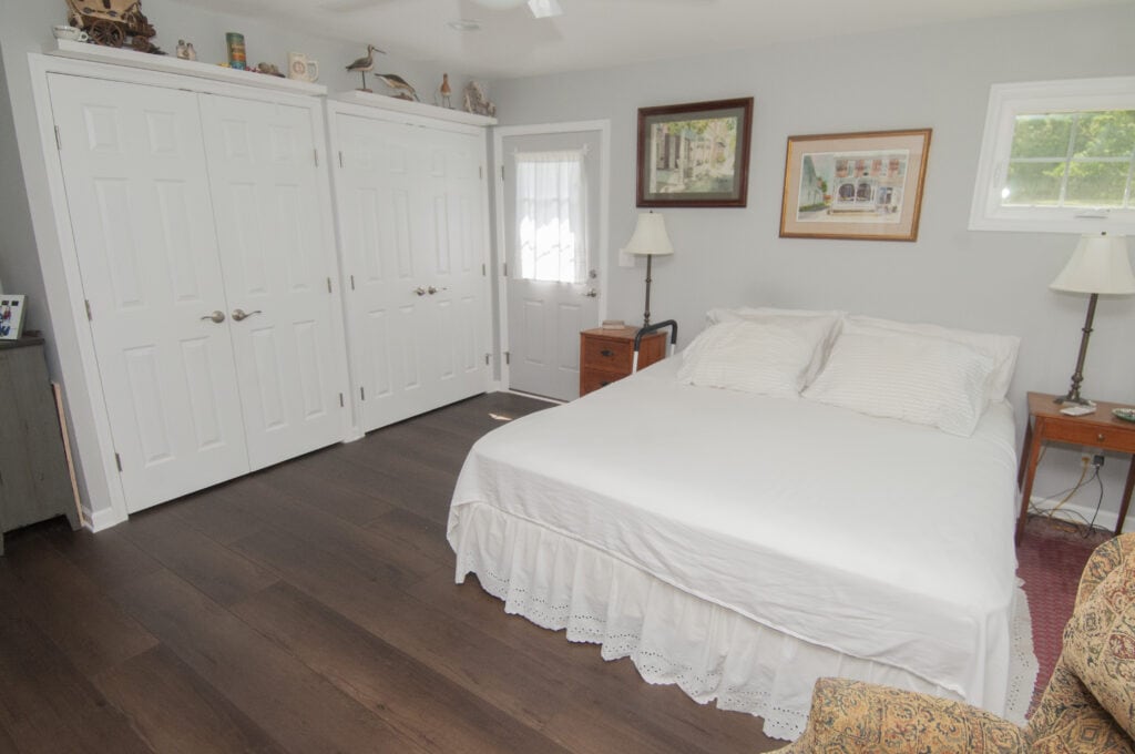 A bright bedroom with dark wood floor, a neatly made bed with white bedding, two nightstands with lamps, double white closets, a door with a sheer curtain, and framed pictures on light gray walls.