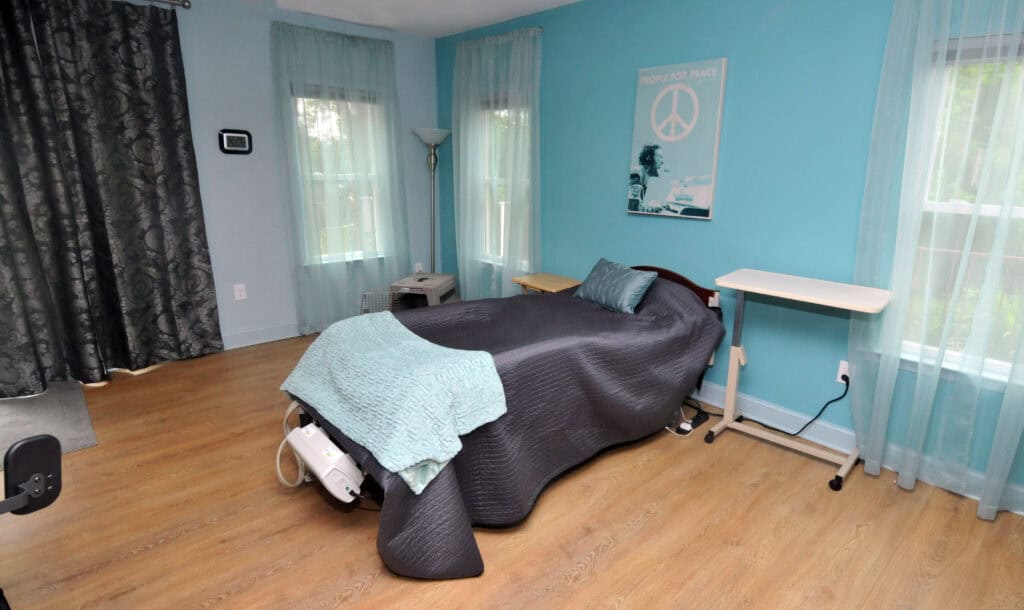 A neatly made bed with a gray cover and teal blanket in a bright, tidy room with blue walls, light wood floors, large windows with sheer curtains, and a peace sign poster on the wall.