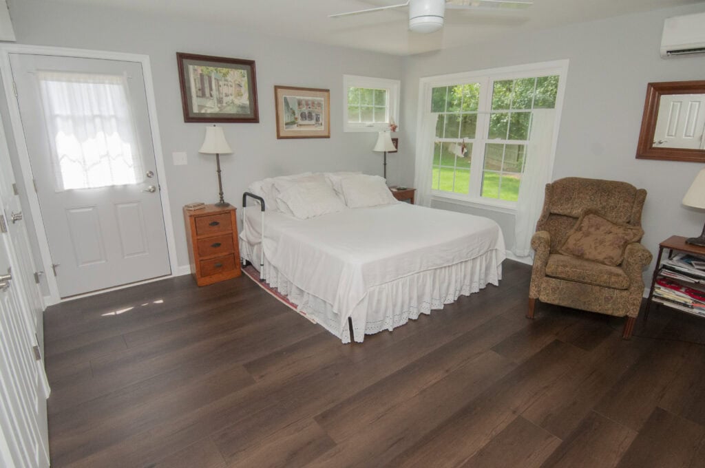 A bright bedroom with a large bed covered in white linens, two wooden nightstands with lamps, an upholstered armchair, framed pictures on light gray walls, and large windows overlooking a green yard.