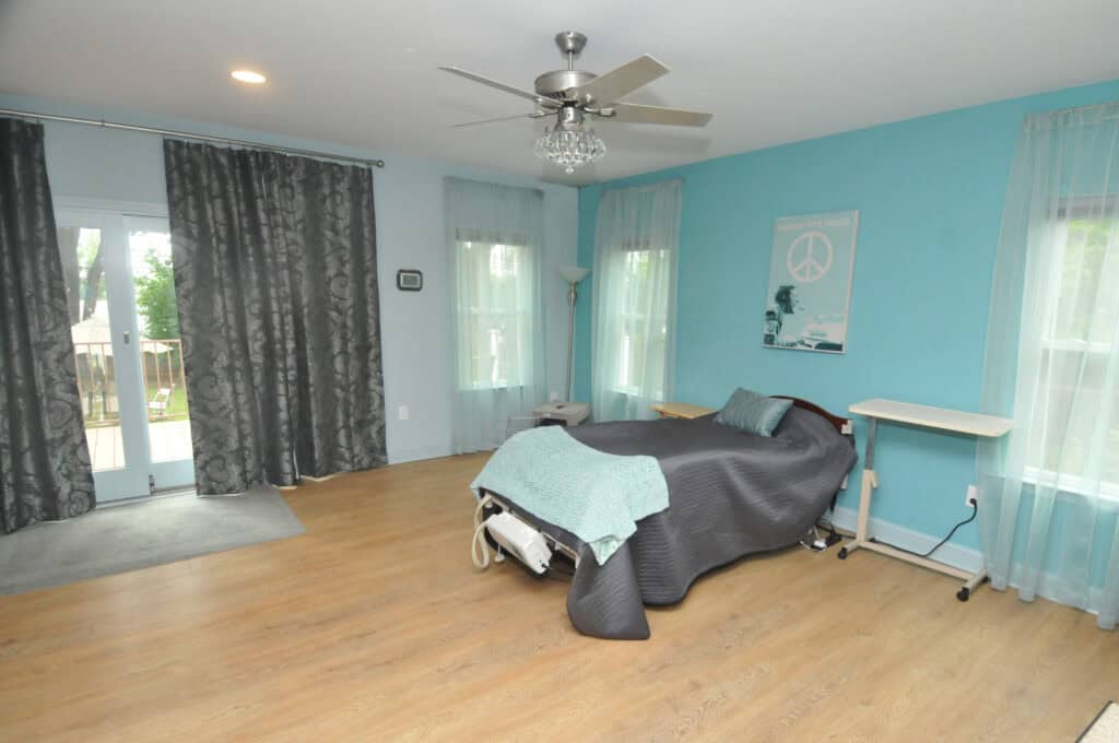 A spacious bedroom with light blue walls, a wooden floor, a twin bed with gray and blue bedding, sheer curtains, a ceiling fan, and a Peace poster on the wall. Theres also a standing lamp and an overbed table.