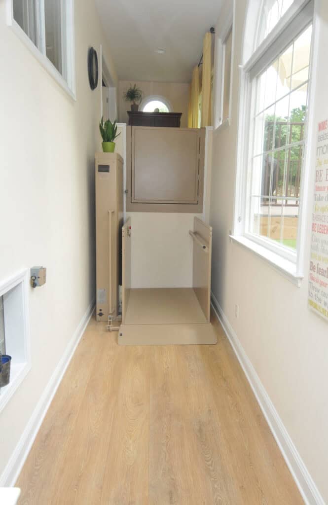 A vertical home lift with its door open is installed in a narrow, brightly lit hallway with light wood flooring and large windows on the right side.