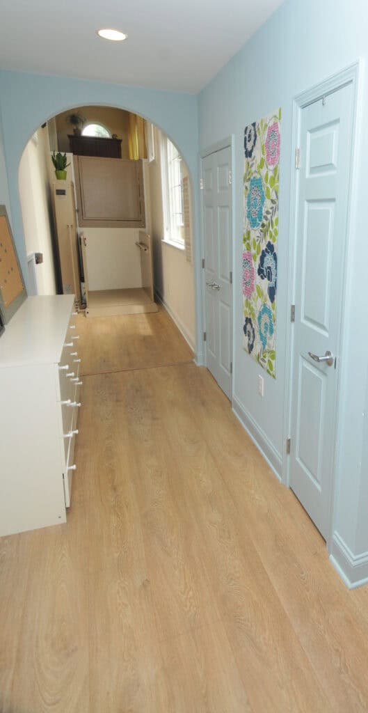A hallway with light wooden floors, pale blue walls, white doors, a white dresser with open drawers, two colorful floral paintings, and an archway leading to another room with windows and plants.