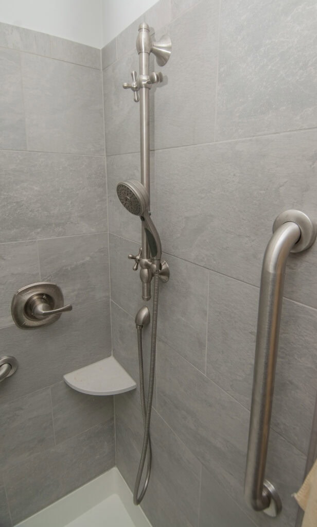 A shower with light gray tiled walls, a metal handheld showerhead on a vertical bar, a wall-mounted grab bar, a water control handle, and a small corner shelf.
