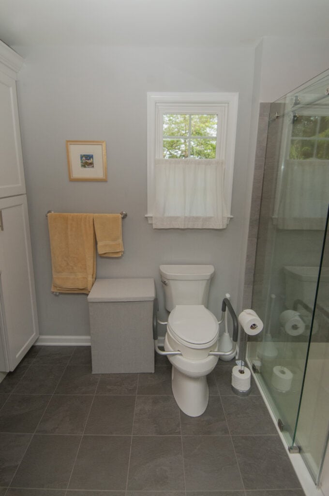 A small, modern bathroom with gray tile flooring, a white toilet with grab bars, a toilet paper holder, a gray storage bench, yellow towels on a rack, a framed picture, a curtained window, and a glass shower door.