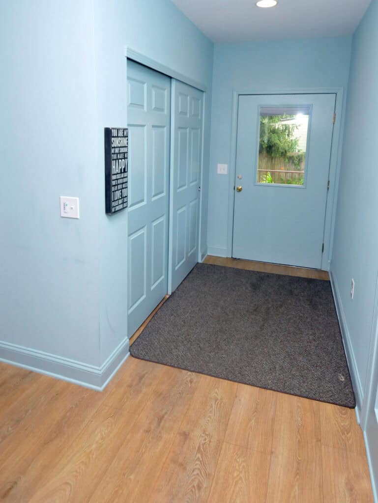 A narrow hallway with light blue walls and doors, wood flooring, a dark gray doormat, and a wall sign with inspirational text. A glass door at the end shows greenery outside.