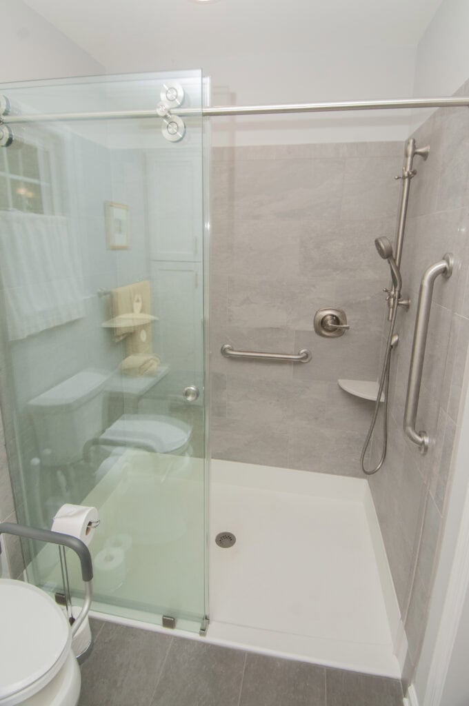 A modern walk-in shower with gray tiles, a glass sliding door, a chrome showerhead, a grab bar, a built-in corner shelf, and a handheld shower wand. A toilet is visible just outside the shower.