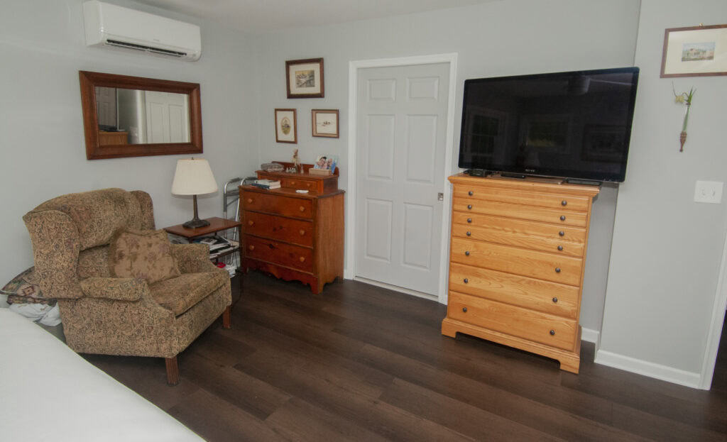A bedroom with dark wood floor, a patterned armchair, a lamp, two wooden dressers, a large flat-screen TV, framed pictures on light gray walls, and a white door in the center.