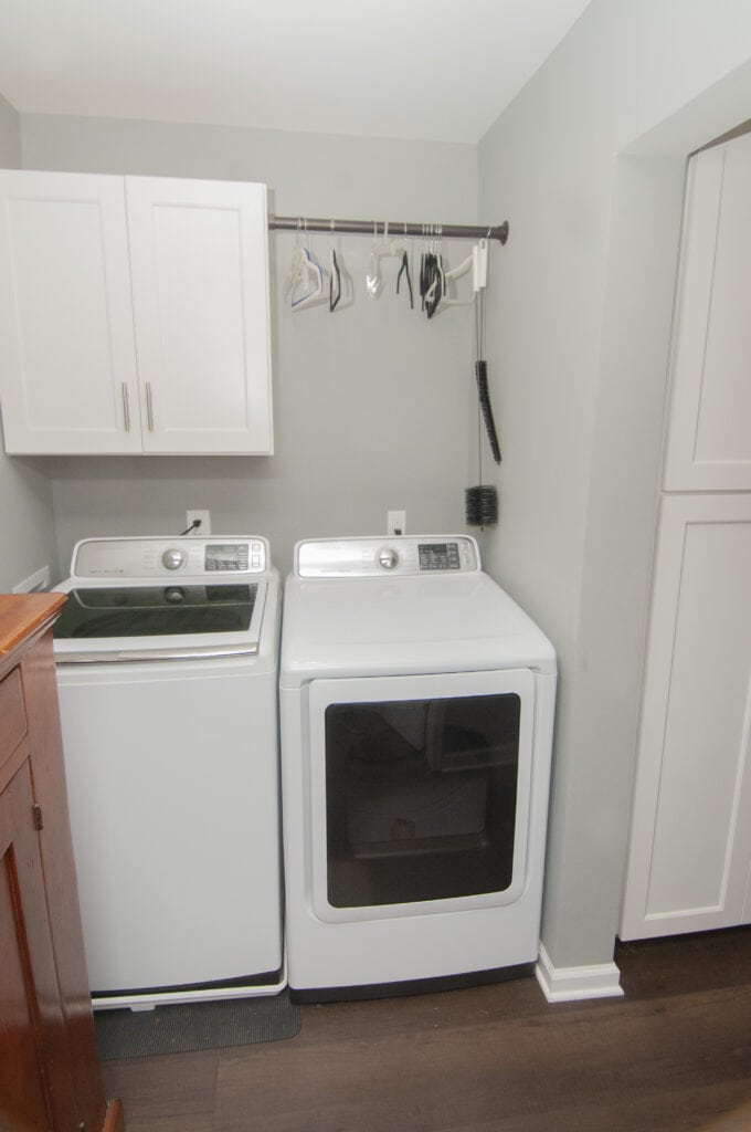 A laundry room with a washing machine and dryer side by side, white cabinets above, cleaning tools and hangers on a rod, and dark wood flooring.