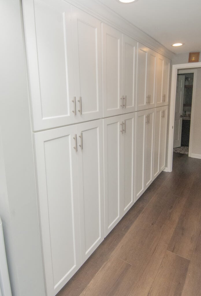A row of tall, white cabinets with silver handles is built along a hallway wall with dark wood flooring and recessed ceiling lights. A doorway is visible at the end of the hallway.