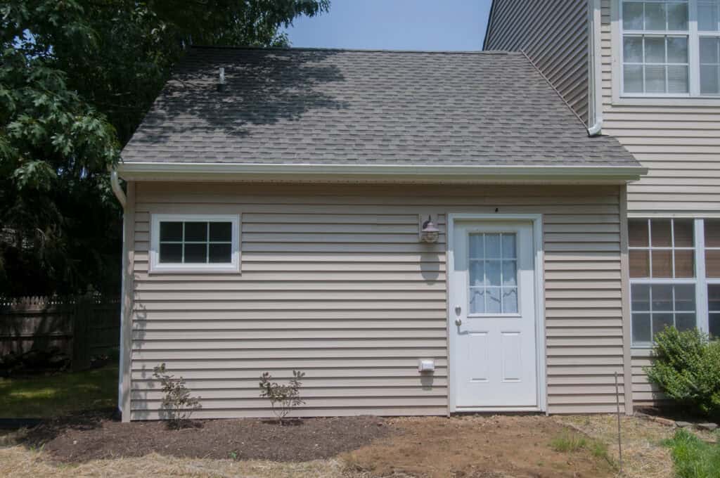 A beige house with horizontal siding, a small window on the left, a white door with a window in the center, and a few small shrubs planted in front. A portion of another window is visible on the right.