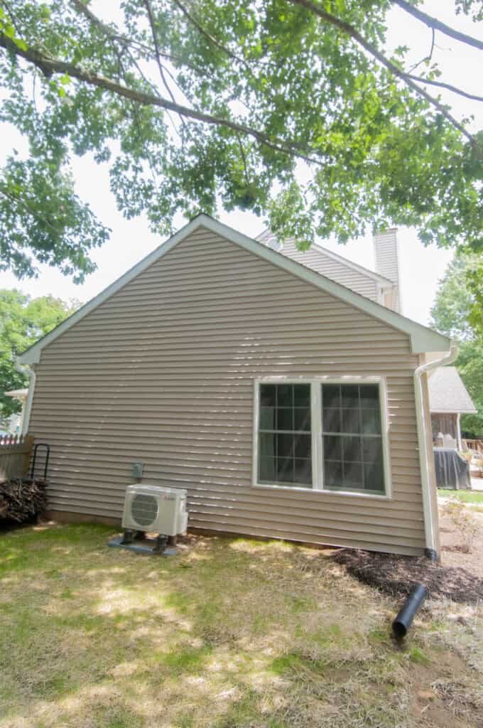 The image shows the exterior side of a beige house with vinyl siding, a large window, and an air conditioning unit on the ground. There are trees overhead and some patchy grass and mulch in the yard.