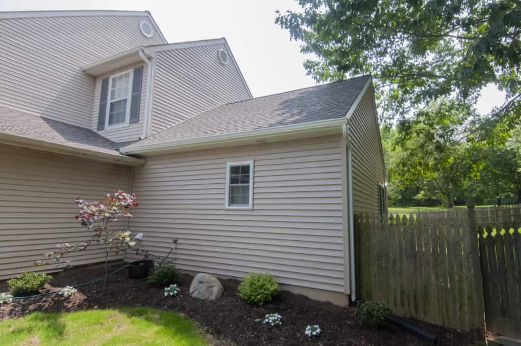 A beige house with vinyl siding, a small window, and a sloped roof is shown next to a wooden fence. In front, there are landscaped flower beds with plants, mulch, and a well-maintained lawn. Trees are visible in the background.