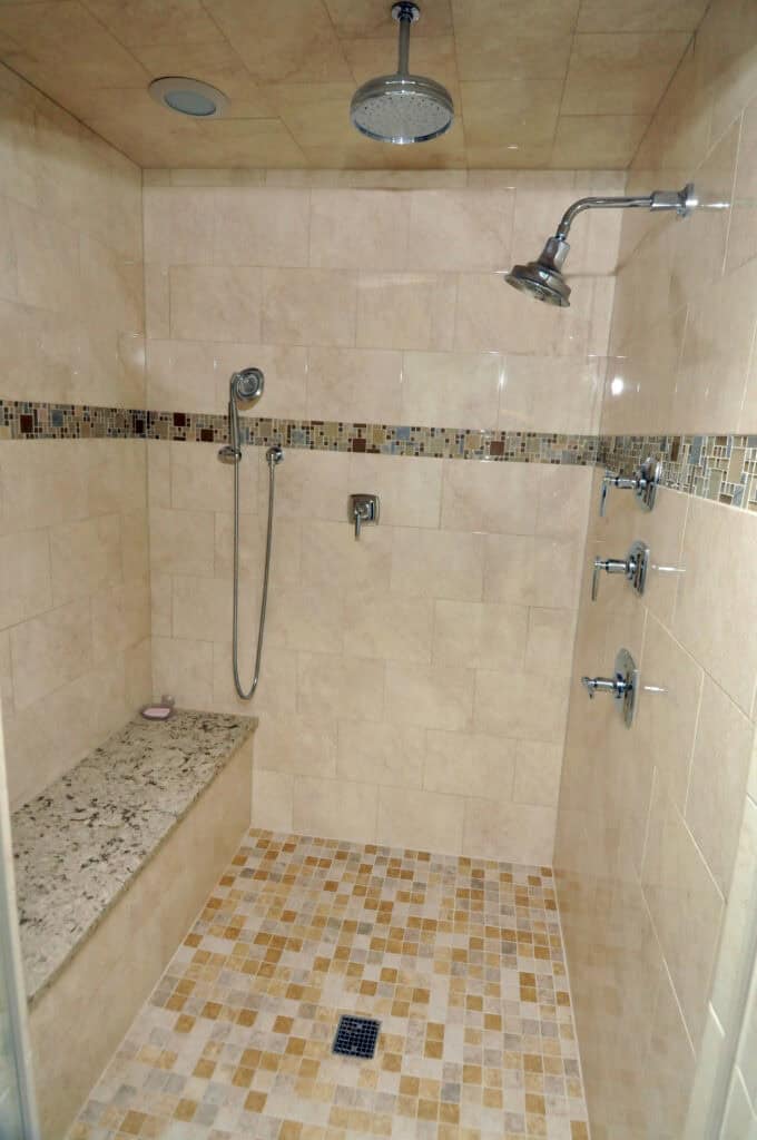 Spacious tiled shower with a built-in bench, rain showerhead on the ceiling, handheld showerhead, multiple wall jets, and decorative tile accent strip. The floor features small square tiles in earth tones.