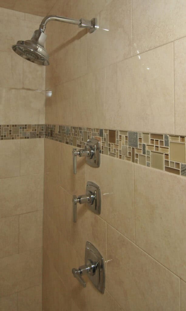 A tiled shower wall with three chrome handles aligned vertically and a showerhead above, featuring beige tiles and a decorative mosaic border.
