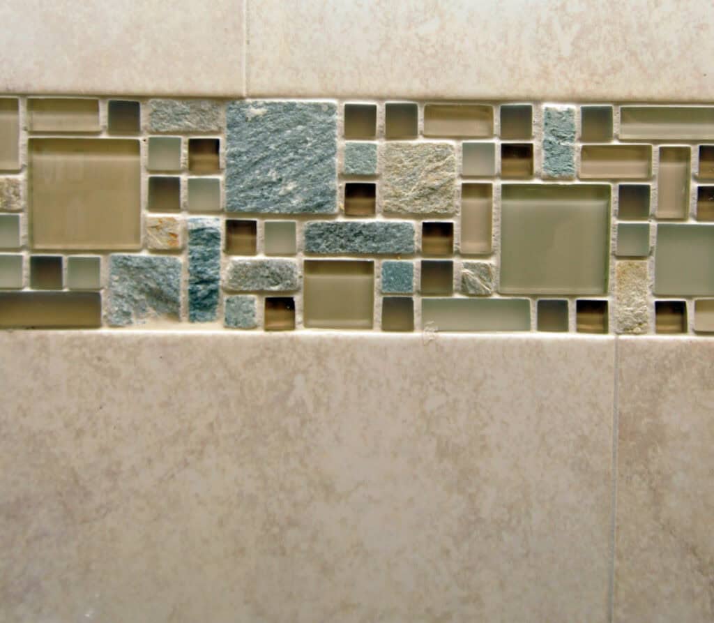 A close-up of a tiled wall featuring a decorative horizontal strip with a mix of small square and rectangular tiles in shades of blue, green, and beige, set within larger beige tiles.