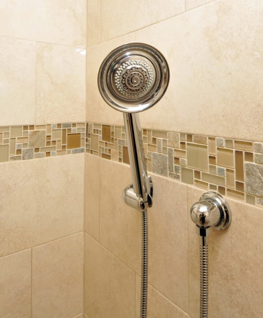 A modern handheld showerhead with a chrome finish is mounted on a tiled shower wall with beige tiles and a decorative mosaic border.