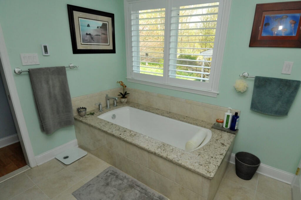 A bathroom with a rectangular soaking tub surrounded by beige tiles, a gray towel on a rack, toiletries by the tub, a window with white shutters, and light green walls decorated with framed photos.