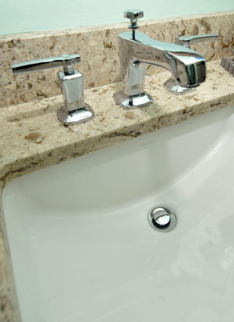 A close-up of a bathroom sink with a polished chrome faucet and handles set in a beige, speckled stone countertop. The white sink is clean, and the drain stopper is closed.