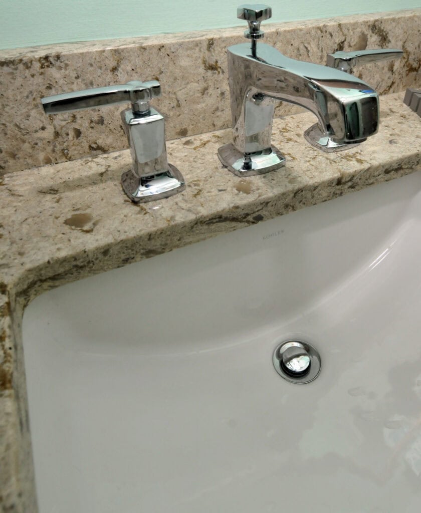 A close-up view of a bathroom sink with a granite countertop, chrome faucet, and handles. The sink is clean and white, with the drain stopper visible at the bottom.