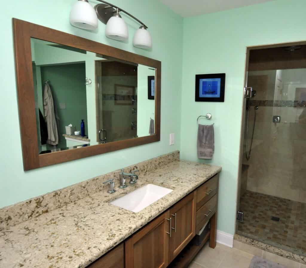 Modern bathroom with a granite countertop, wood vanity, large mirror, three-light fixture, towel ring with gray towel, and a glass shower with tiled walls and floor. Walls are painted a light mint green.