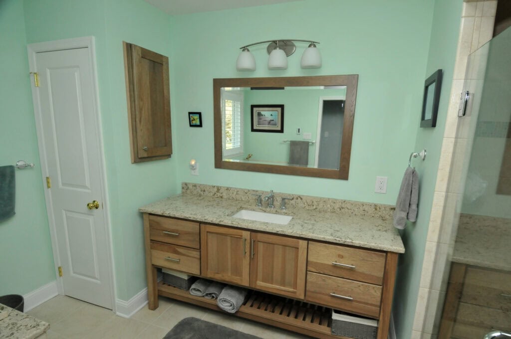 A bathroom with mint green walls, a wooden vanity with a marble countertop, a rectangular mirror, three-light fixture, and neatly rolled towels on the lower shelf. A door and a towel hanging on the wall are visible.