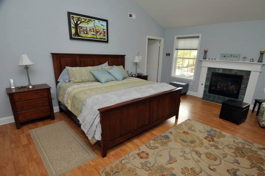 A spacious bedroom with a wooden bed, patterned bedding, two nightstands, a rug, a fireplace, and a window letting in daylight. A colorful painting hangs above the bed. The room has light-colored walls and wood flooring.