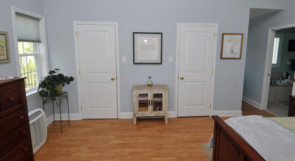 A bedroom scene with two white doors on a light blue wall. A small cabinet with a plant and decor sits between the doors. Wood floor, framed art, and furniture are visible. Natural light comes from a window on the left.