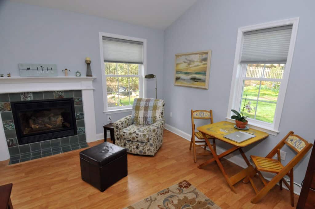 A cozy living room with a fireplace, patterned armchair, small wooden table with a laptop and plant, folding chairs, hardwood floor, and two windows letting in natural light, overlooking a green backyard.