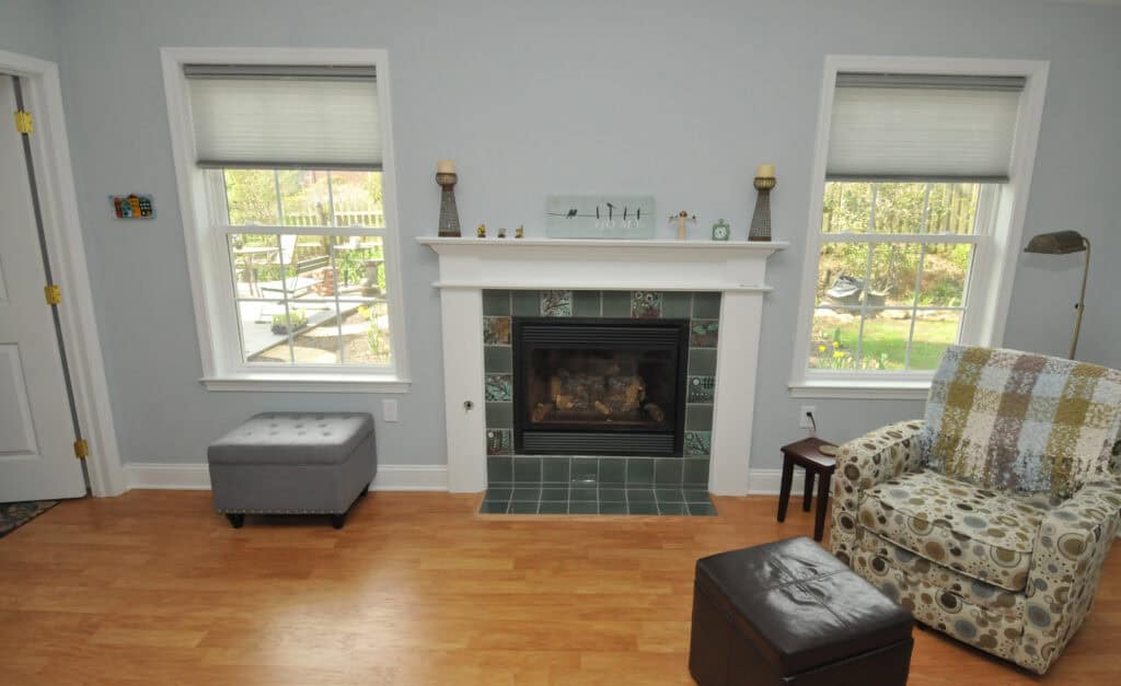 A cozy living room with a fireplace centered between two windows, an armchair with a patchwork quilt, a small brown ottoman, a gray upholstered bench, and hardwood floors. Greenery is visible outside the windows.