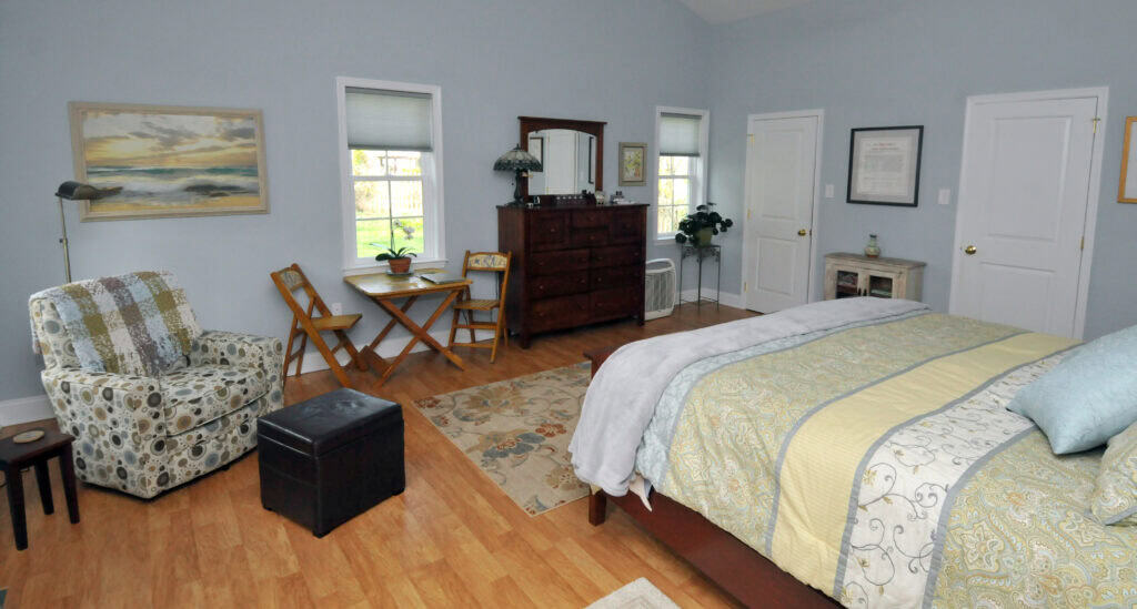 A cozy bedroom with light blue walls, wooden floor, a patterned armchair, a bed with yellow and gray bedding, a dresser with a mirror, a small table with two chairs, and several windows letting in natural light.
