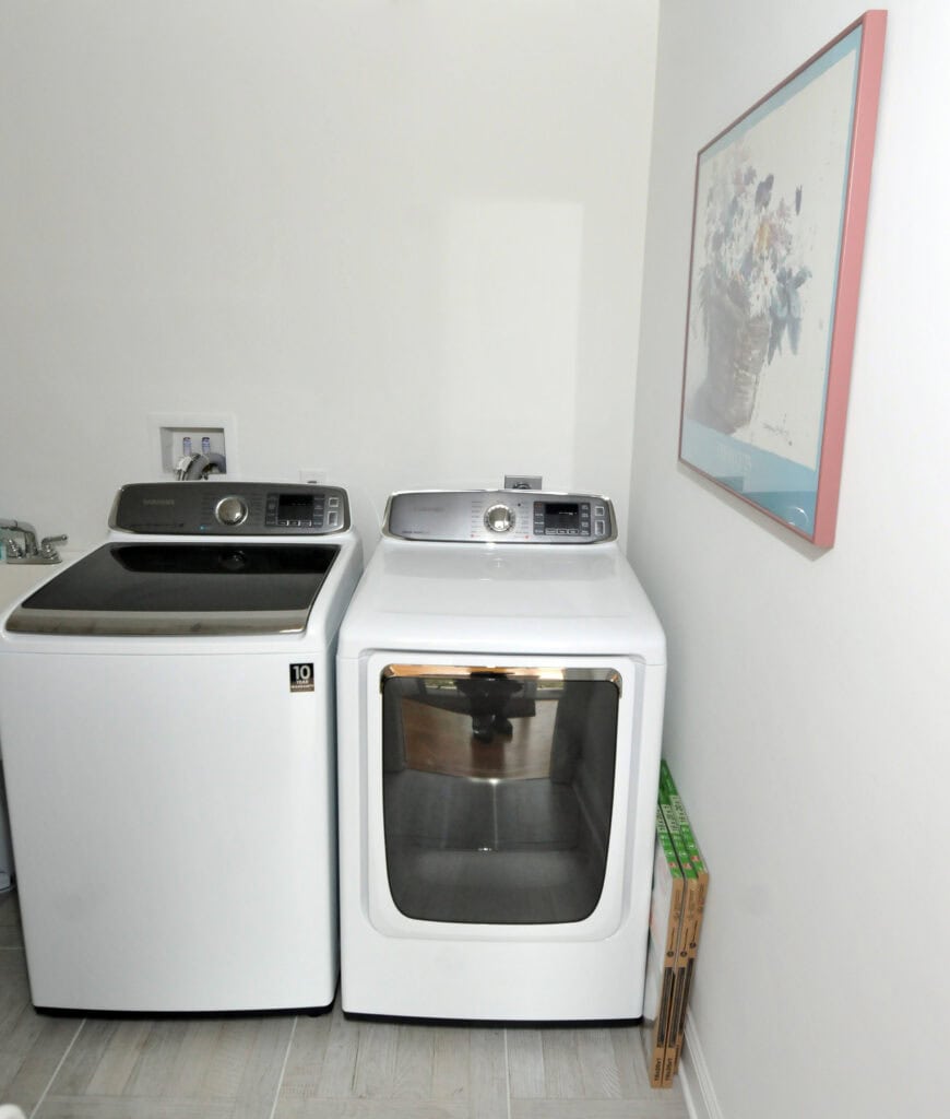 A modern laundry room with a top-load washing machine and a front-load dryer side by side, a floral painting on the wall, and a few narrow boxes leaning against the wall.