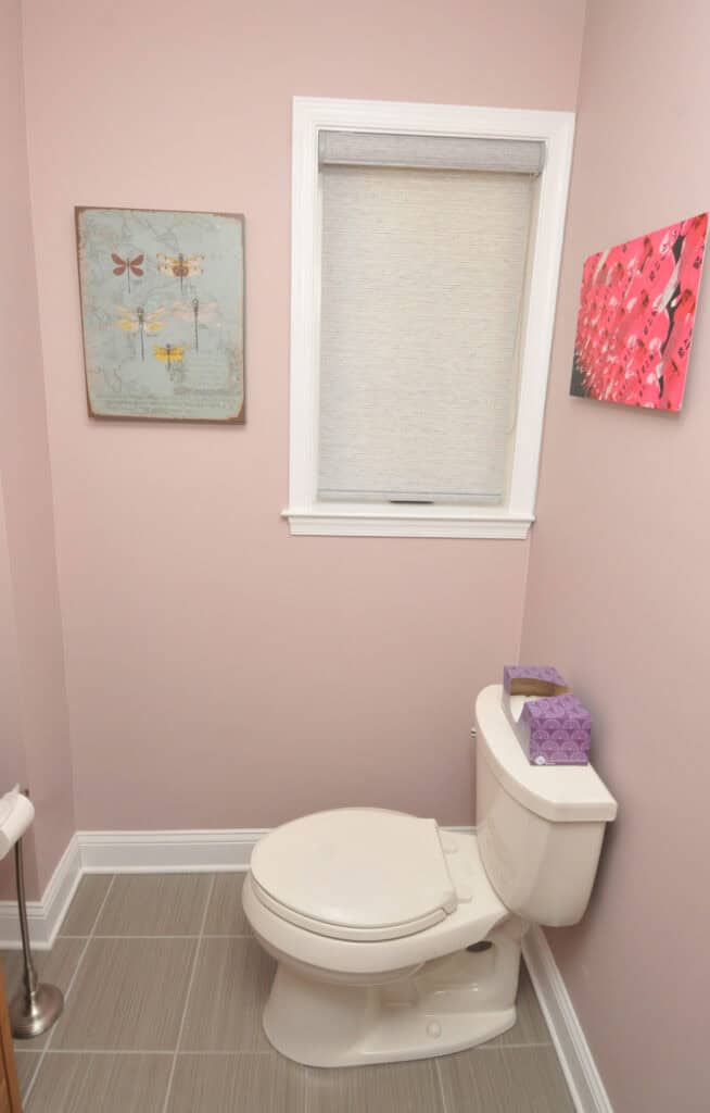 A small bathroom with light pink walls, a white toilet, a box of purple tissues on the tank, wall art featuring dragonflies and flowers, a window with a shade, and light gray tile floor.